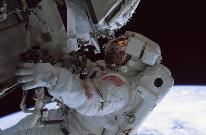 An astronaut in a spacesuit is performing a spacewalk with the Earth and space visible in the background.