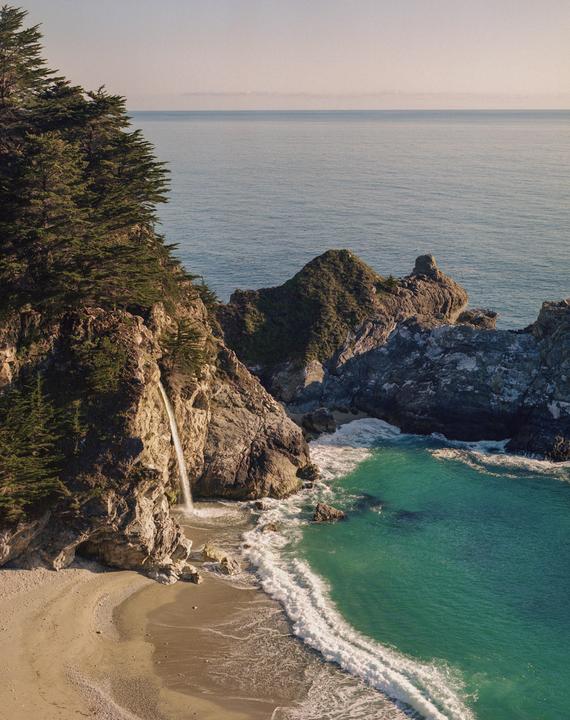 A waterfall cascades onto a sandy beach next to the ocean, surrounded by rocky cliffs and green trees.