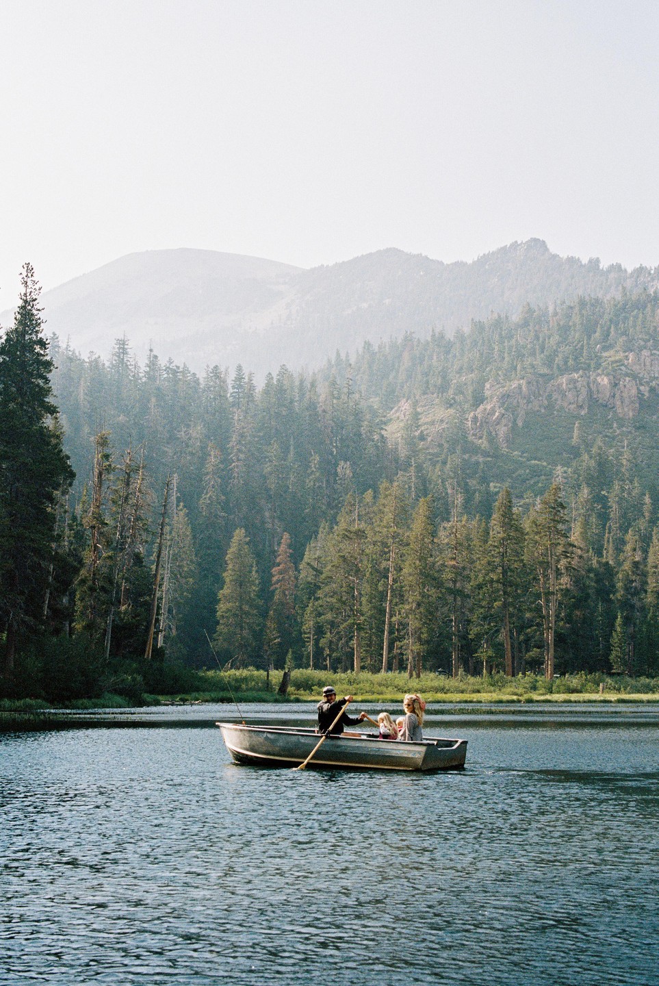 A man rows a boat with a woman and child on a lake surrounded by a forest and mountains.