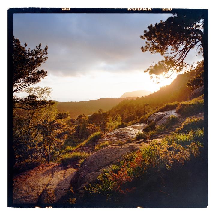 The sun sets over a mountainous landscape with trees and rocky terrain.