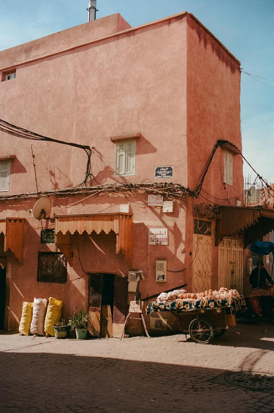 A pink building with white window shutters and a street vendor's cart filled with bread sits on a sunny street.