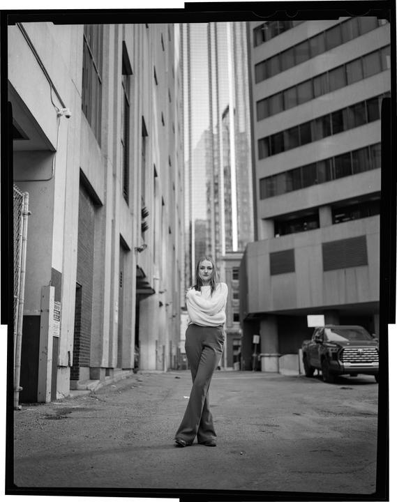 A woman stands in an alley with her arms crossed, looking at the viewer.