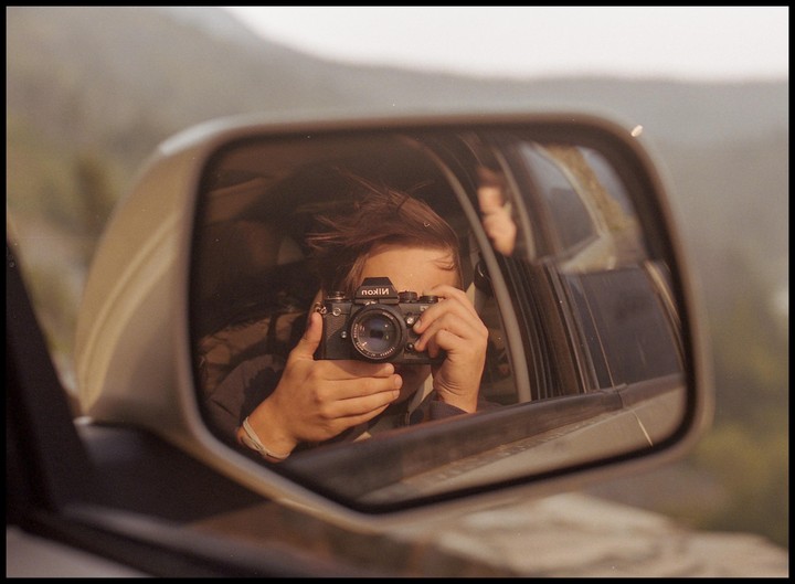 A person holds a camera up to their face, reflected in a car's side mirror.