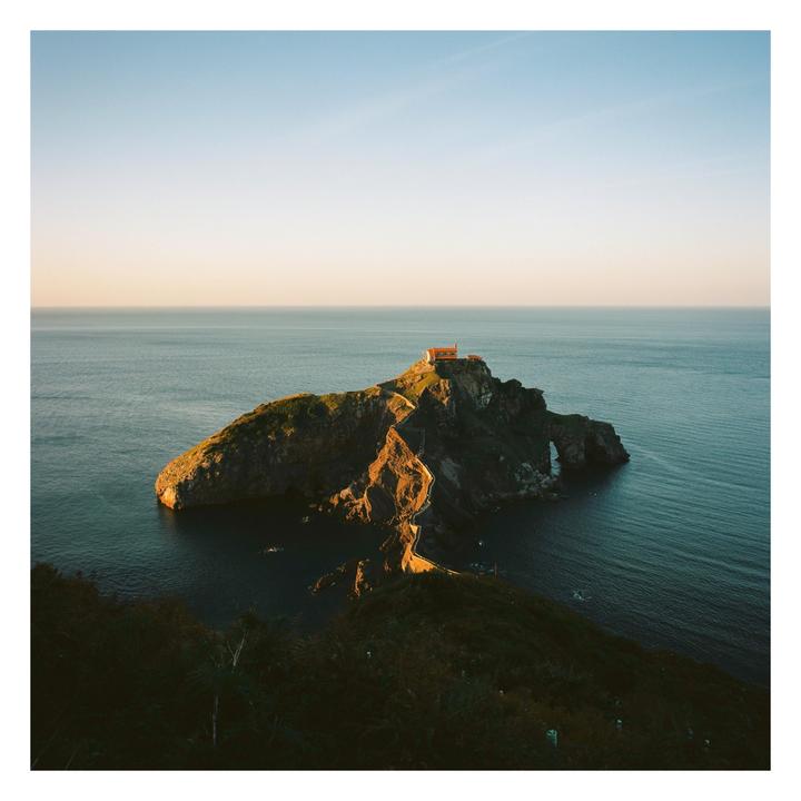 A small building sits atop a rocky island connected to the mainland by a winding path, surrounded by the sea under a clear sky.