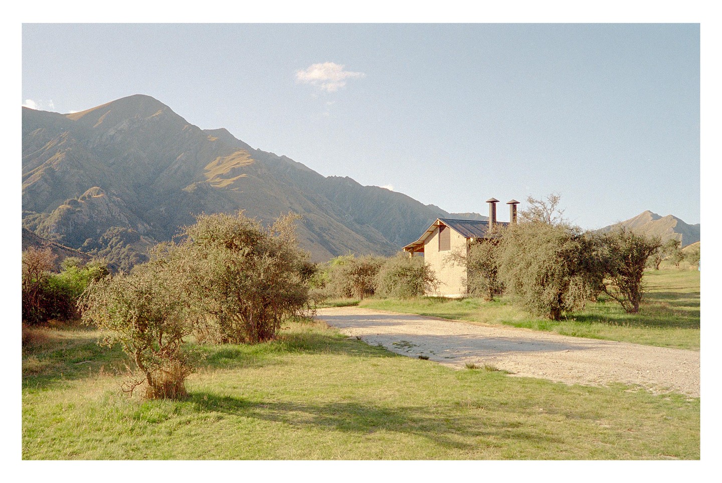 A cabin with two chimneys sits among trees and a dirt road, with mountains in the background under a clear sky.