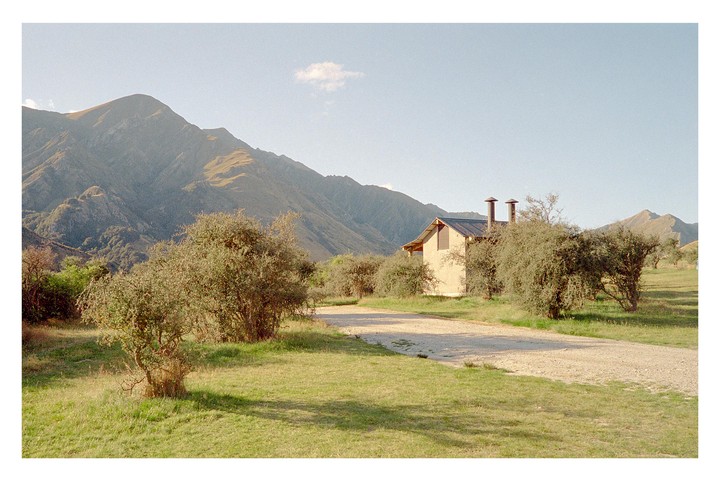 A cabin with two chimneys sits among trees and a dirt road, with mountains in the background under a clear sky.