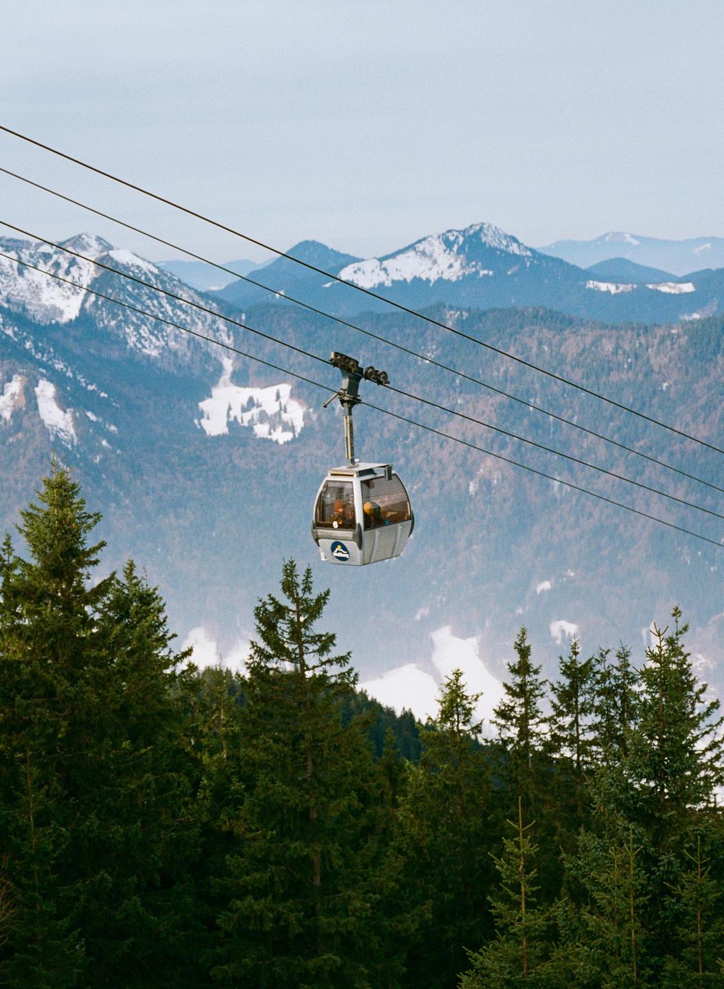 A gondola with people inside is suspended by cables above a forest with snow-capped mountains in the background.