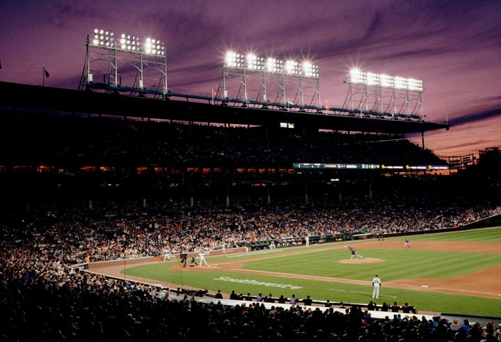 A baseball game is being played in a stadium at dusk with bright stadium lights illuminating the field.