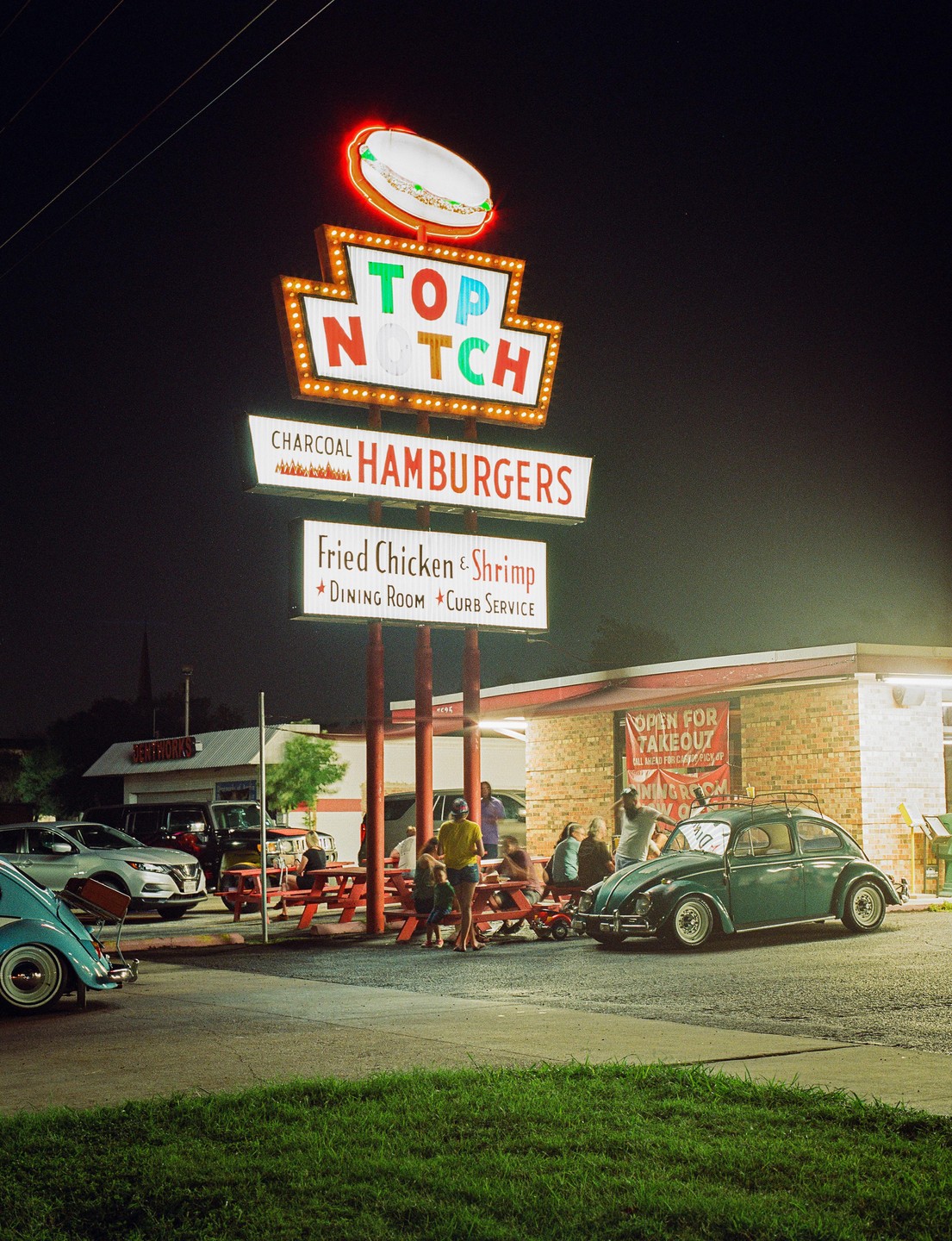 A neon sign for a hamburger restaurant stands tall over a parking lot with people and cars at night.