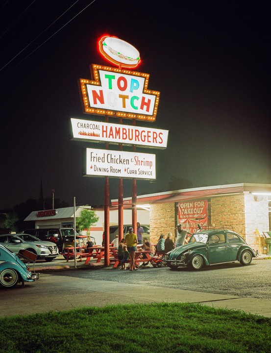 A neon sign for a hamburger restaurant stands tall over a parking lot with people and cars at night.