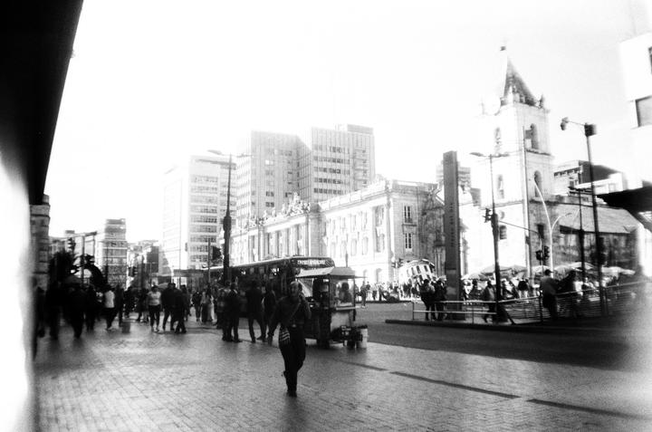 A busy street in Bogota is filled with people, a bus, and a food cart, with a church and other buildings in the background.