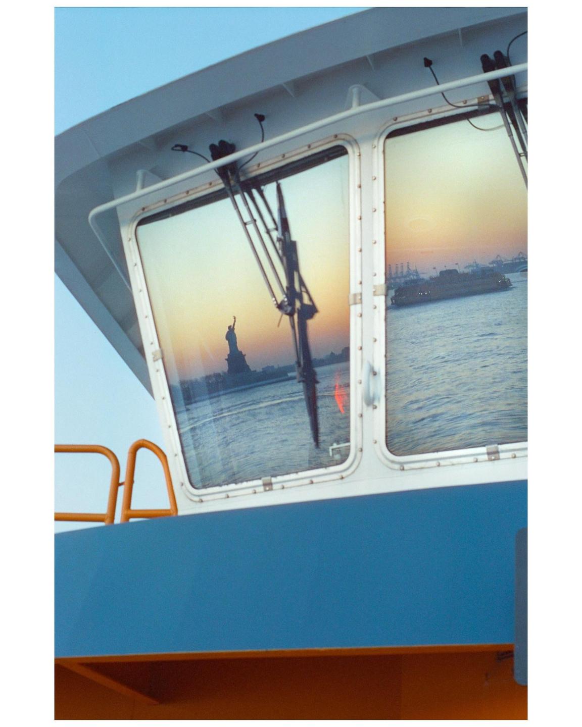 The Statue of Liberty and a ferry are reflected in the windows of a boat at sunset.