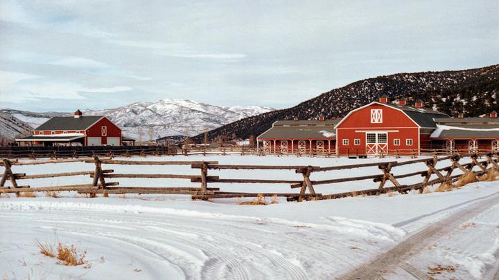 Two red barns with white trim stand behind a wooden fence in a snowy field with mountains in the background.