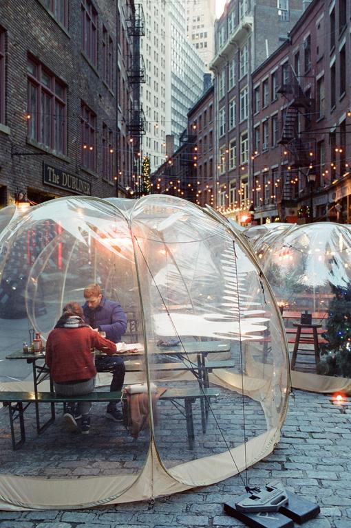 A couple dines inside a clear orb on a cobblestone street lined with buildings and string lights.
