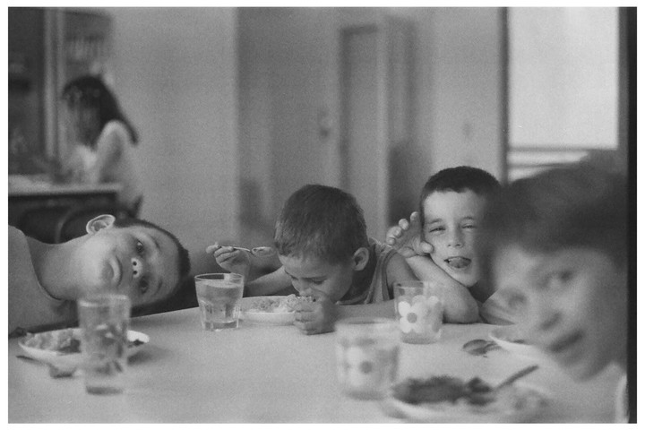 Three boys are sitting at a table, one of them is eating, another is sticking out his tongue, and the third is looking at the camera.