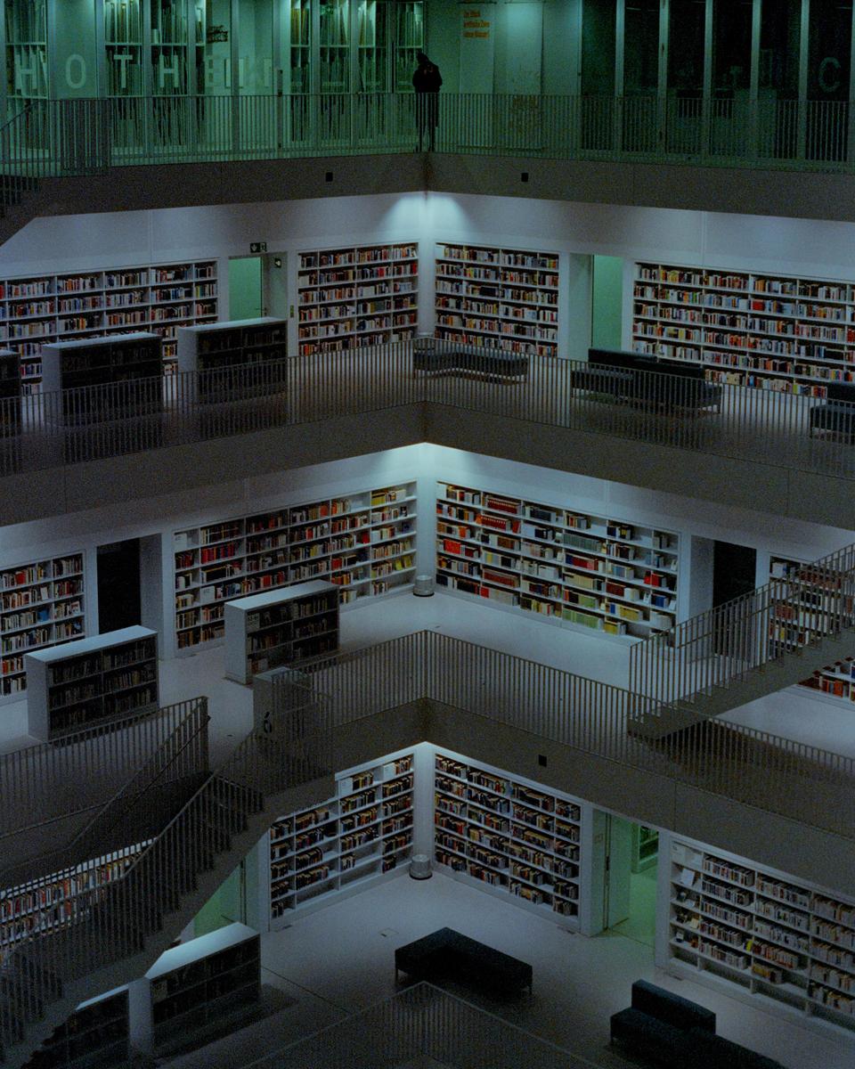 A person stands on an upper level overlooking a multi-story library with many bookshelves and staircases.