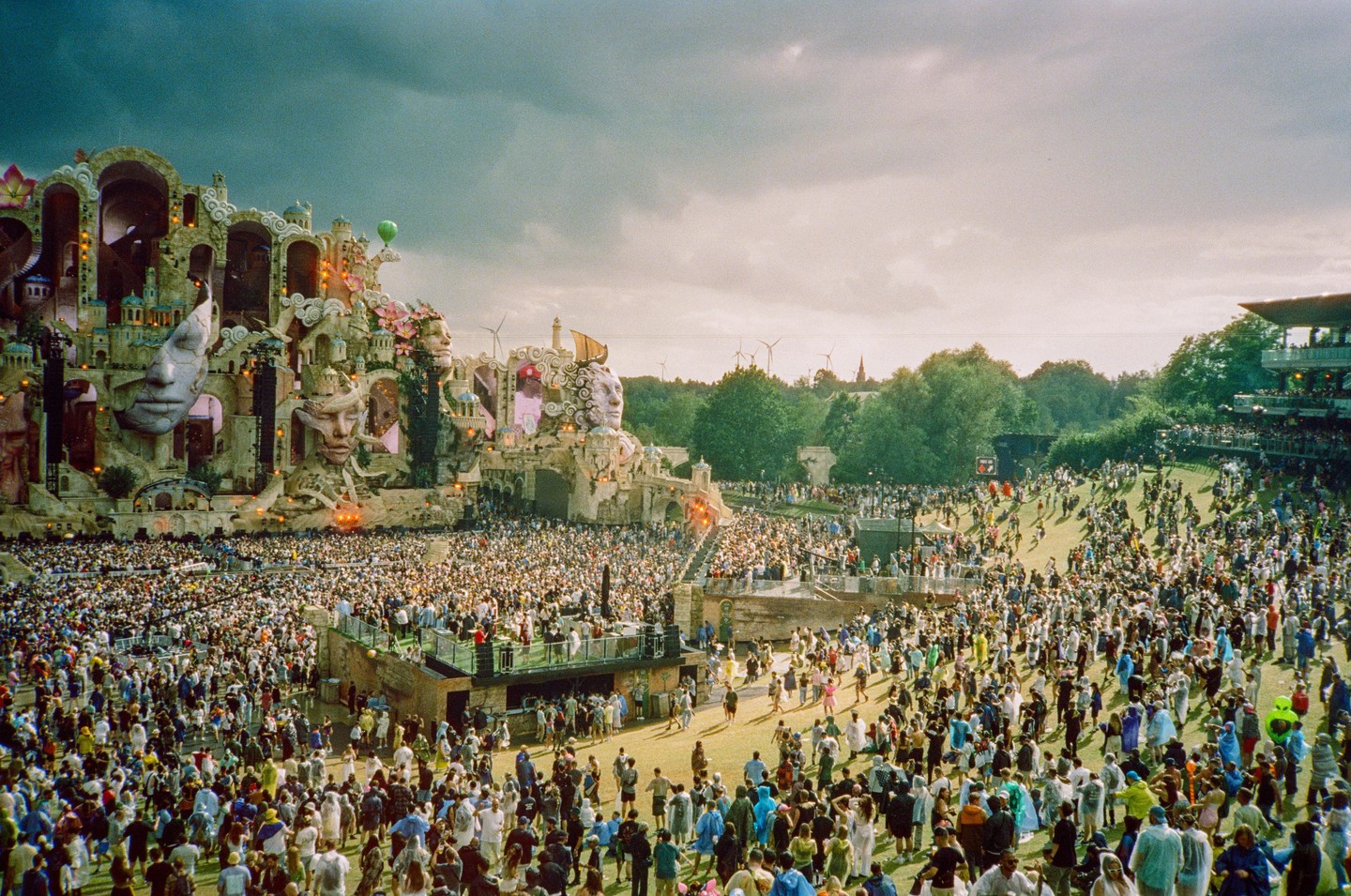 A large crowd of people gathers in front of an elaborate stage with sculpted faces and architectural details under a cloudy sky.