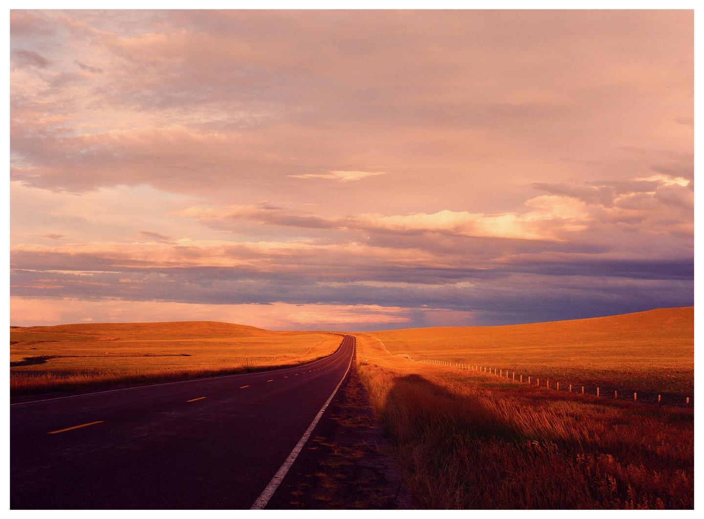 A long road stretches through a field under a dramatic sky at sunset.