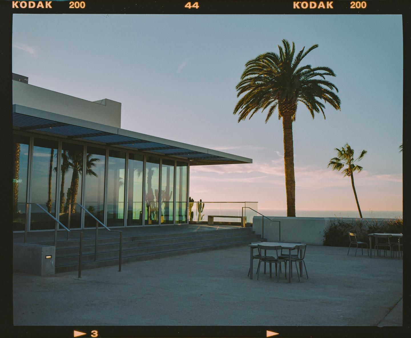 A modern building with large glass windows stands next to palm trees and outdoor seating at sunset.