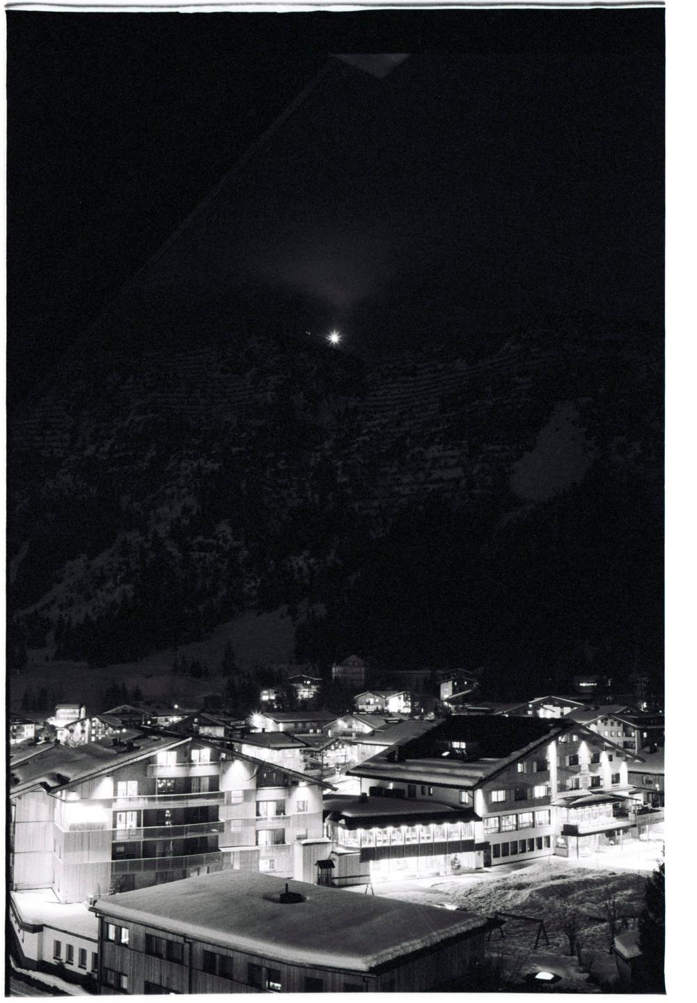 A small town with snow-covered roofs is nestled at the base of a dark, snow-dusted mountain at night.
