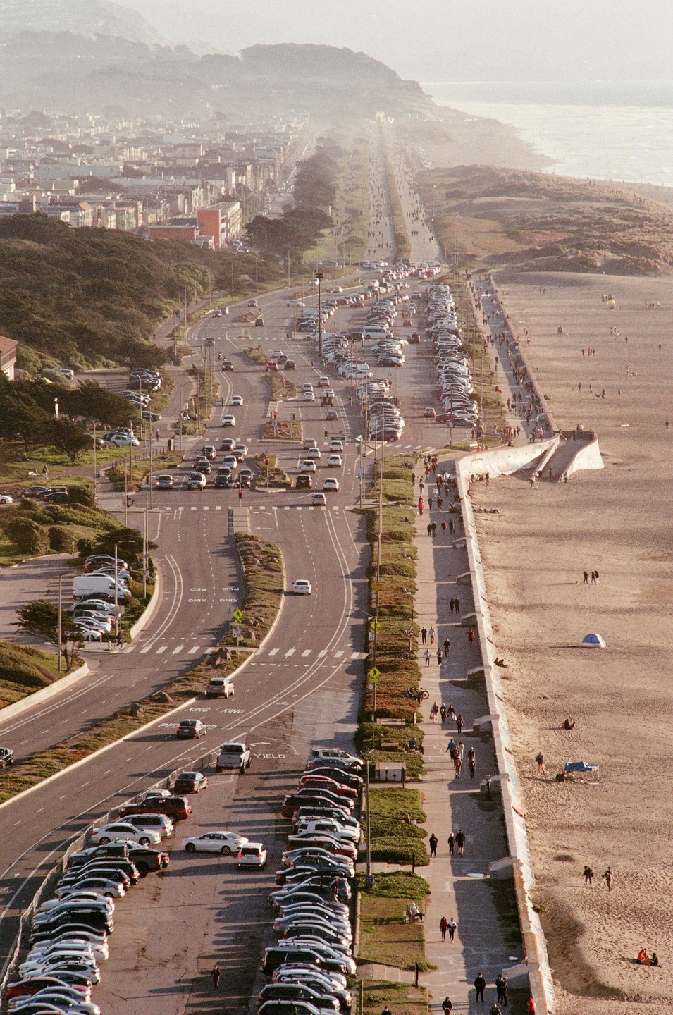 An aerial view shows a highway running parallel to a beach with cars parked along the road and people walking on the sand.