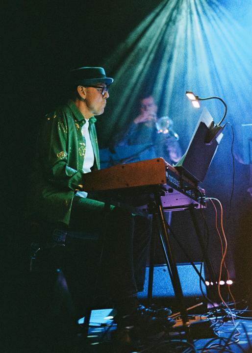 A man in a green shirt and hat plays a keyboard on a stage with blue stage lighting.