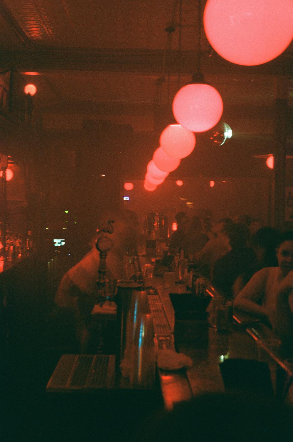 A long bar is lit by a row of red globe lights hanging from the ceiling, with people sitting and standing along its length.