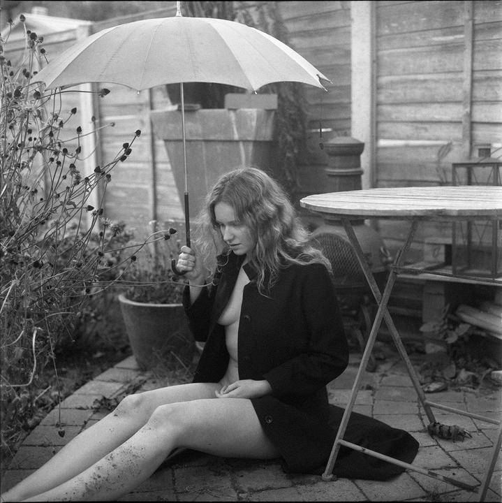A woman sits on a brick patio holding an umbrella, wearing an open coat.