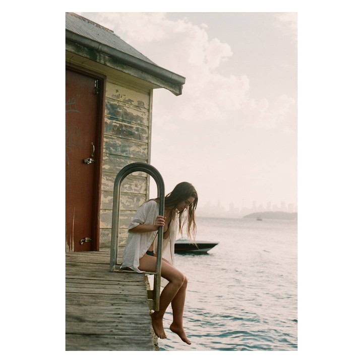 A woman sits on a wooden dock next to a small building, with a boat and city skyline in the background.