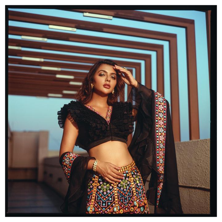 A woman in a black and colorful embroidered outfit poses with her hand on her forehead.