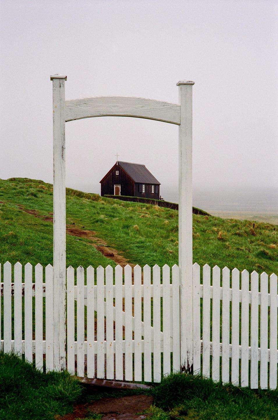 A white picket fence and gate frame a small, dark church on a grassy hill under a hazy sky.