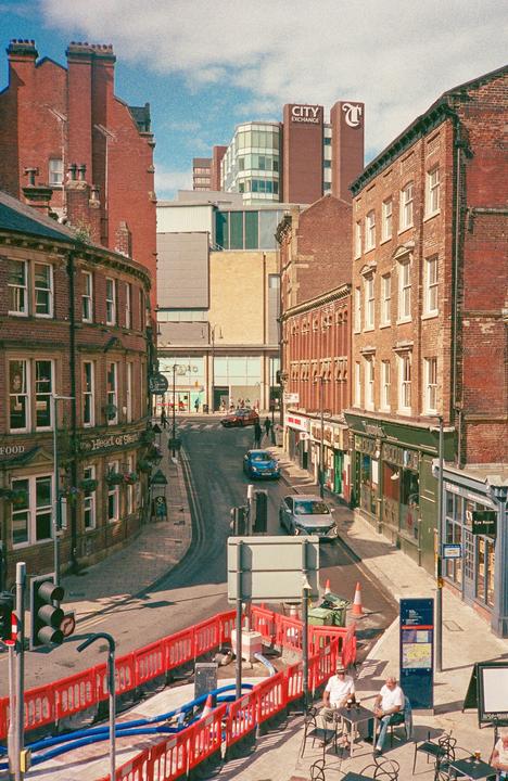 A street in Leeds, England, is lined with brick buildings and cars, with two men sitting at a table in the foreground.
