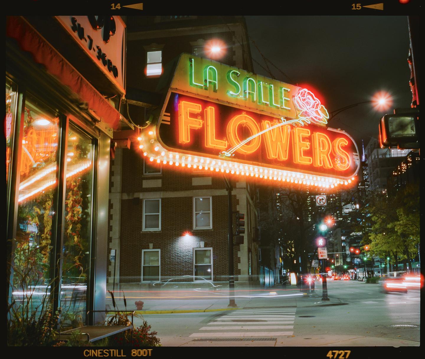 A neon sign for La Salle Flowers glows brightly at night above a street with light trails from passing vehicles.