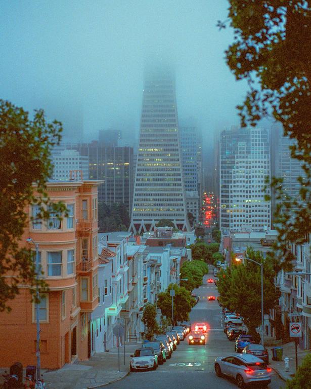 A foggy city street with parked cars and buildings on either side leads to a tall skyscraper partially obscured by fog.