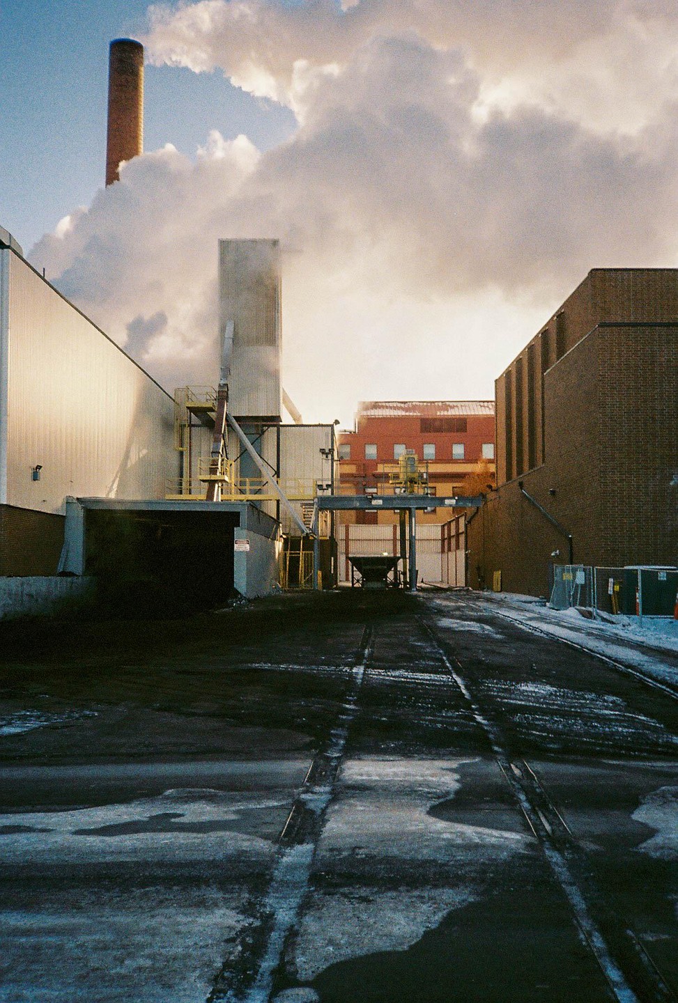 A factory with a tall smokestack emits a large plume of smoke into the sky, with a snowy ground in the foreground.