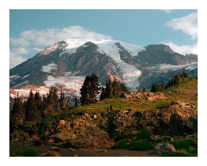 A snow-capped mountain rises above a forested hillside under a blue sky with clouds.