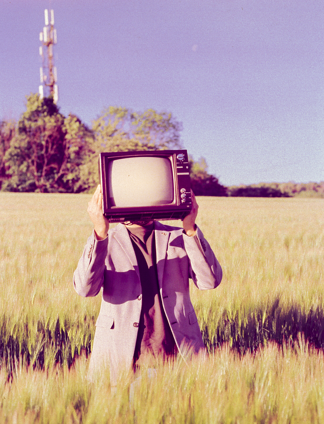 A man in a field holds a television in front of his face, with a communication tower in the background.