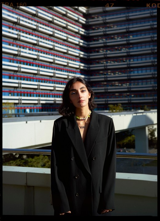 A woman wearing a black blazer and colorful necklaces stands in front of a building with red and white stripes.