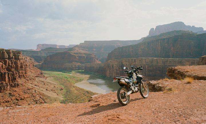 A motorcycle is parked on a dirt path overlooking a river winding through a canyon.