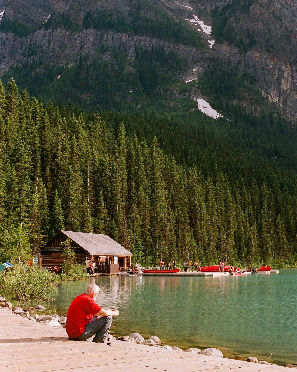 A man in a red shirt sits on a wooden dock by a lake with a boathouse and forest in the background.