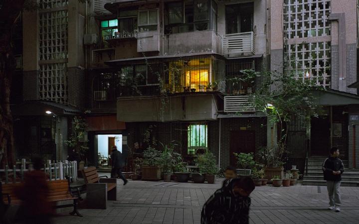 A street at night is lined with buildings and potted plants, with people walking and a bright yellow light emanating from a window.
