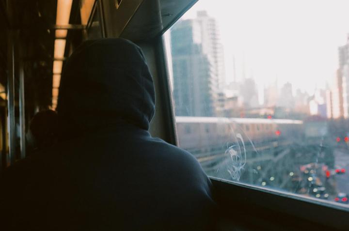 A person in a hoodie looks out a train window at a city skyline and street below.