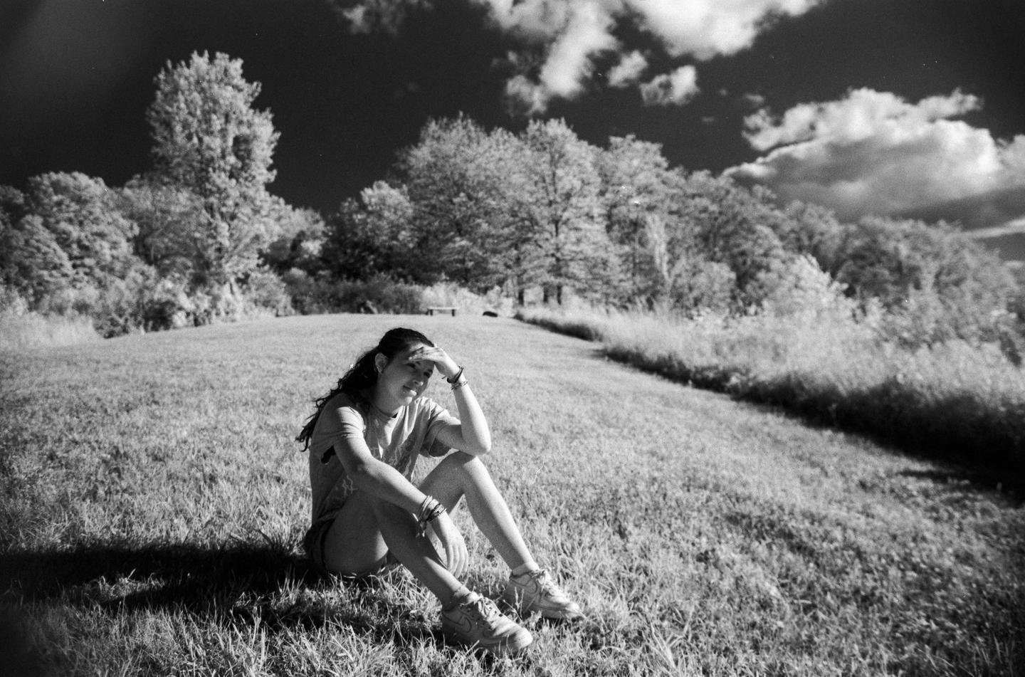 A woman sits on a grassy hill with trees and clouds in the background.