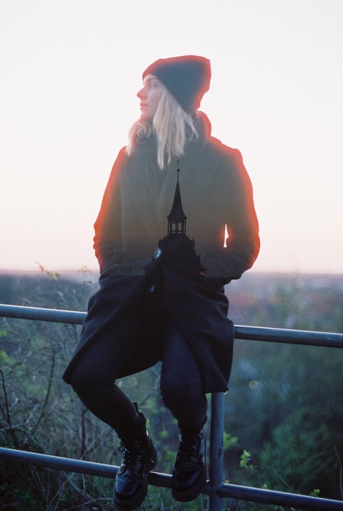 A woman in a beanie and coat sits on a railing with a church steeple double exposed over her body.