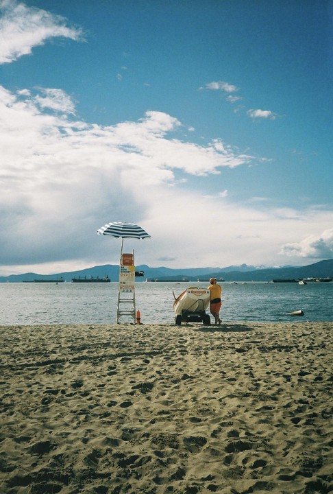 A lifeguard stands next to a boat on a sandy beach with a striped umbrella and a lifeguard chair.