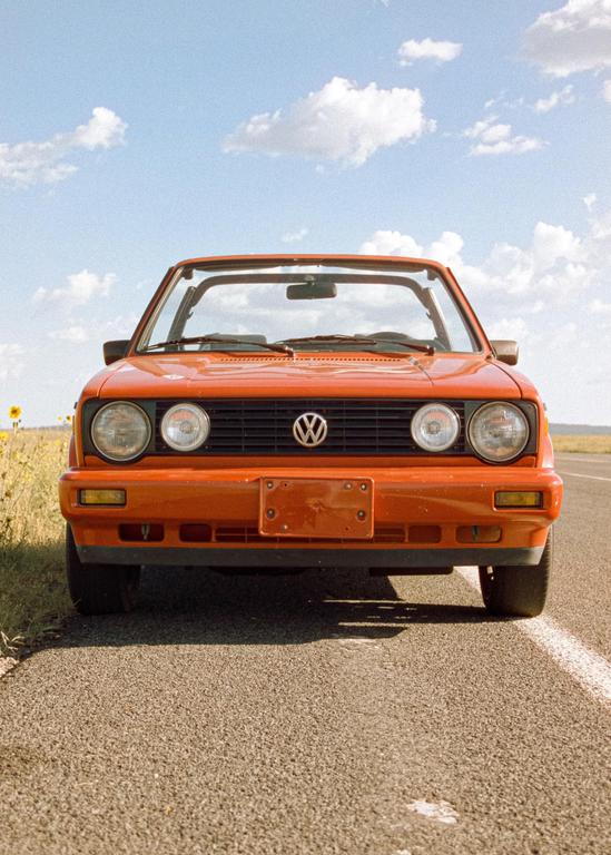 An orange convertible car is parked on the side of a road with a field of sunflowers and a blue sky with clouds in the background.