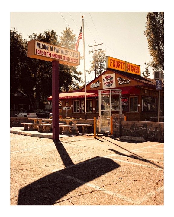 A Frosty Burger restaurant with a large sign and an American flag stands next to a parking lot with long shadows.