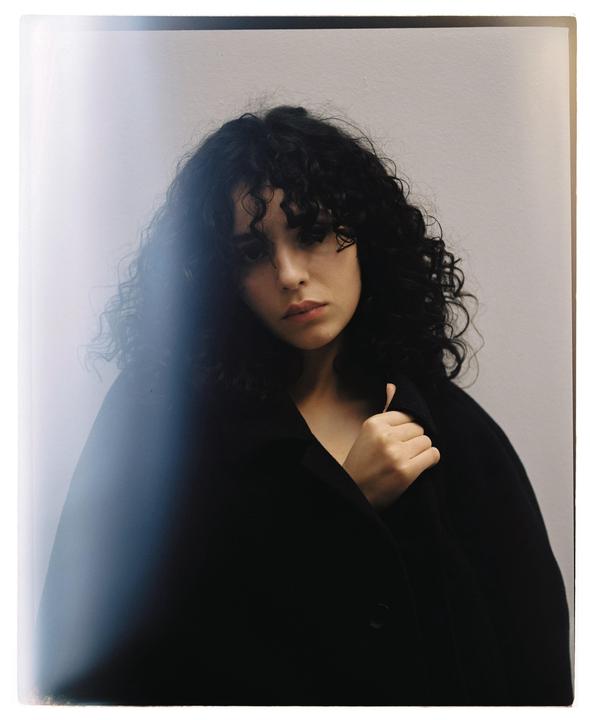 A woman with dark curly hair wears a black coat and looks directly at the viewer.