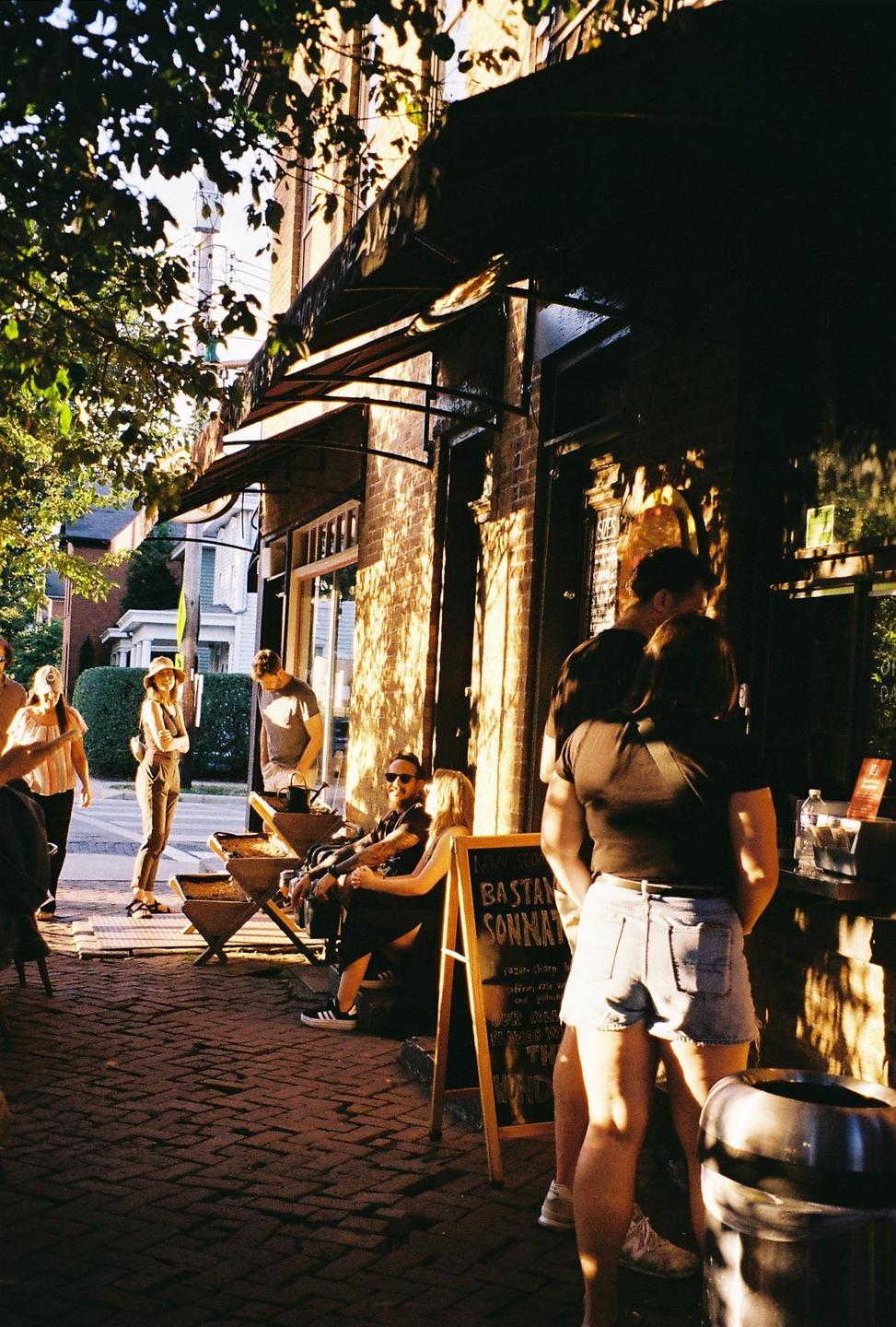 People are gathered on a brick sidewalk outside a building with an awning during golden hour.