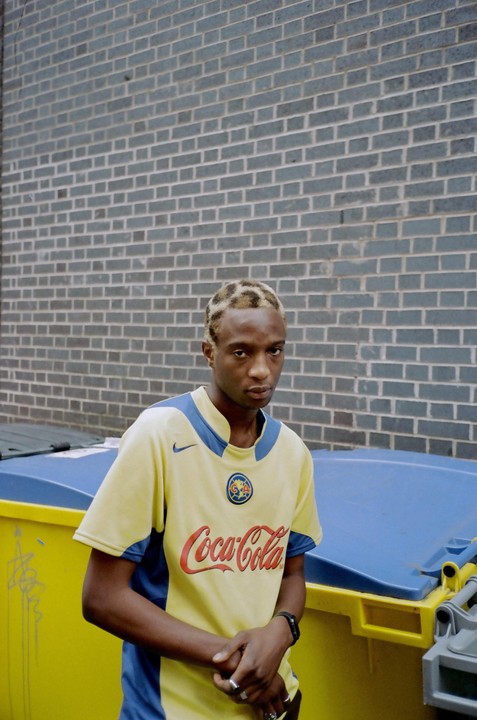 A man with bleached hair and a leopard print pattern stands in front of a brick wall and yellow bins.
